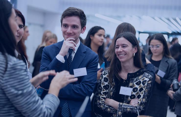 A group of people engaging in conversation at a networking event, with one man and one woman in the foreground smiling and listening attentively.