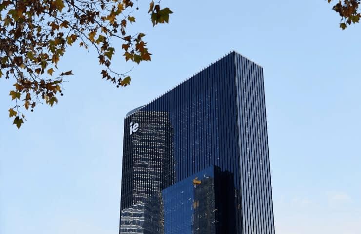 Modern skyscraper with a blue glass facade and the logo 'ie' visible, set against a clear blue sky with autumn leaves in the foreground.