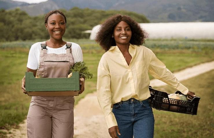 Two women, one in overalls and the other in a yellow blouse, smiling while holding boxes of fresh produce in a field.
