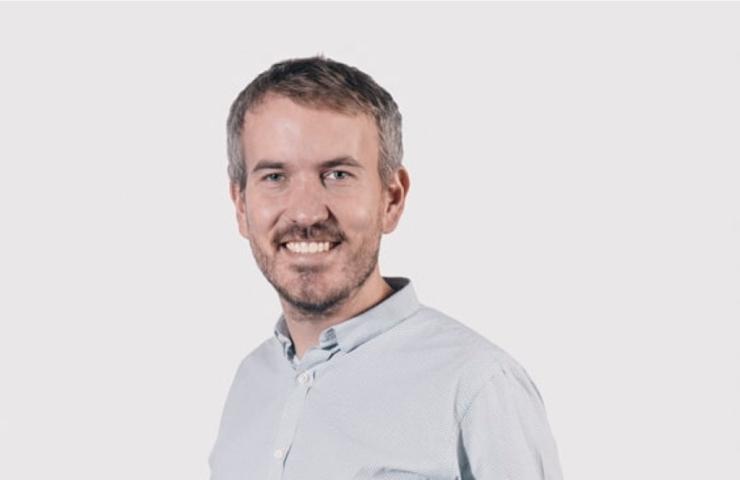 A smiling man wearing a light blue shirt against a white background.