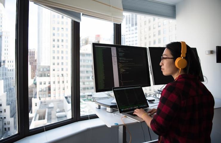 A woman wearing headphones is working on a laptop in front of large windows overlooking a cityscape.