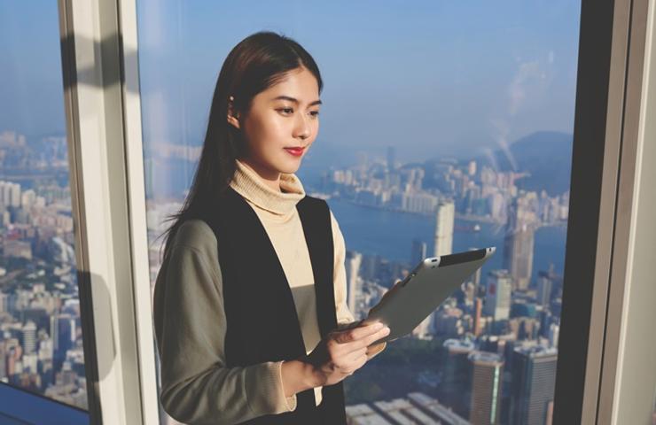 A woman with long hair is holding a tablet while looking out at the city skyline from a tall building.
