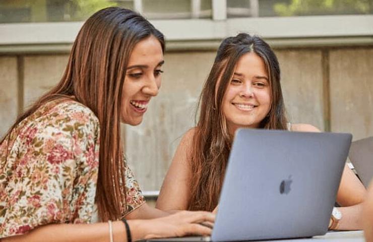 Two women smiling and looking at a laptop outdoors.