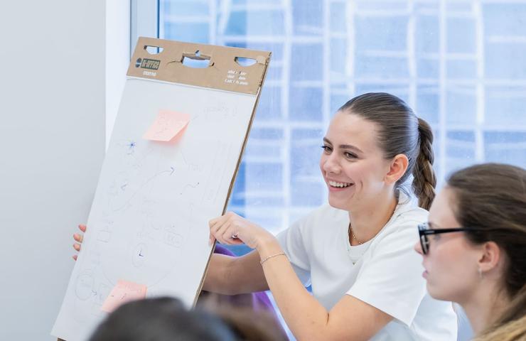 A woman is smiling while presenting a whiteboard with notes.