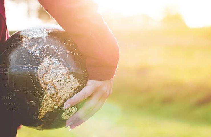 A person holding a small globe in their hands in the sunlight with a natural landscape in the background.