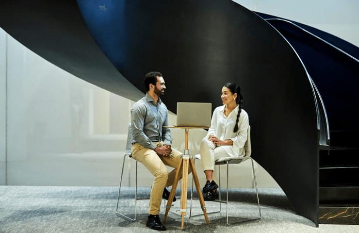 Two people, a man and a woman, having a conversation while sitting on stools near a modern spiral staircase and a laptop on a small table.