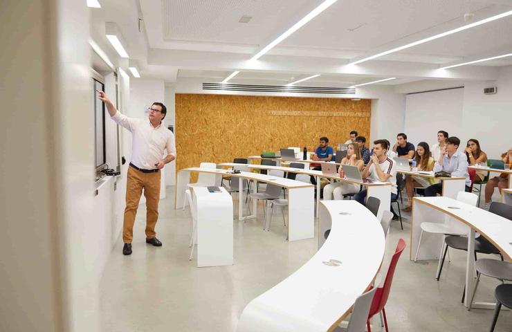 A teacher is explaining in a modern classroom with students seated at curved desks.