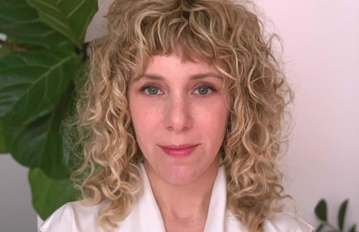 Portrait of a woman with curly blonde hair and a white shirt, standing in front of a green plant.