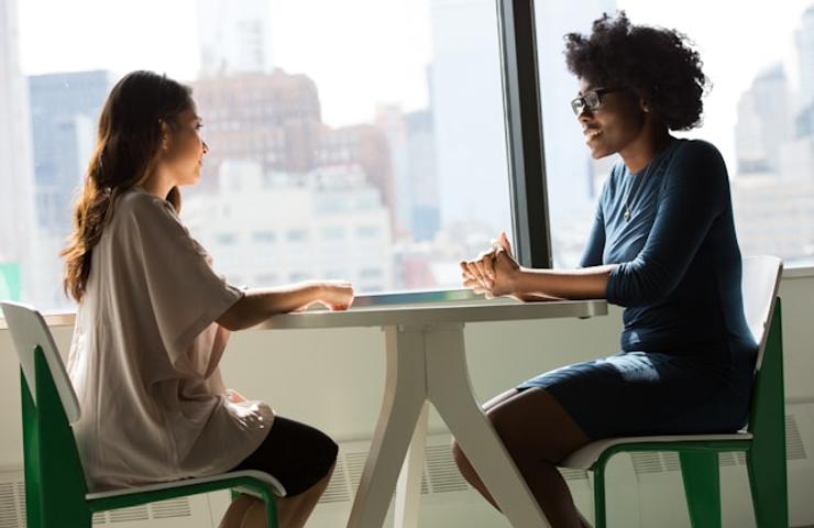 Two women are engaged in a conversation at a table by a large window with a city view.