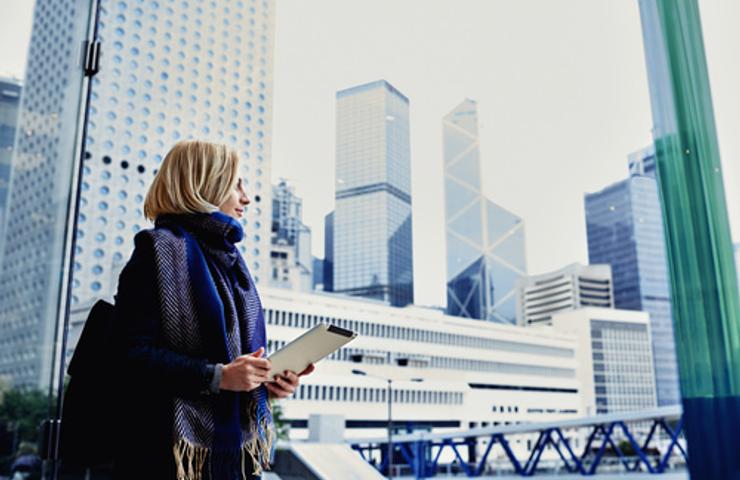 A woman stands in a cityscape, holding a notepad and looking thoughtfully at the skyscrapers.