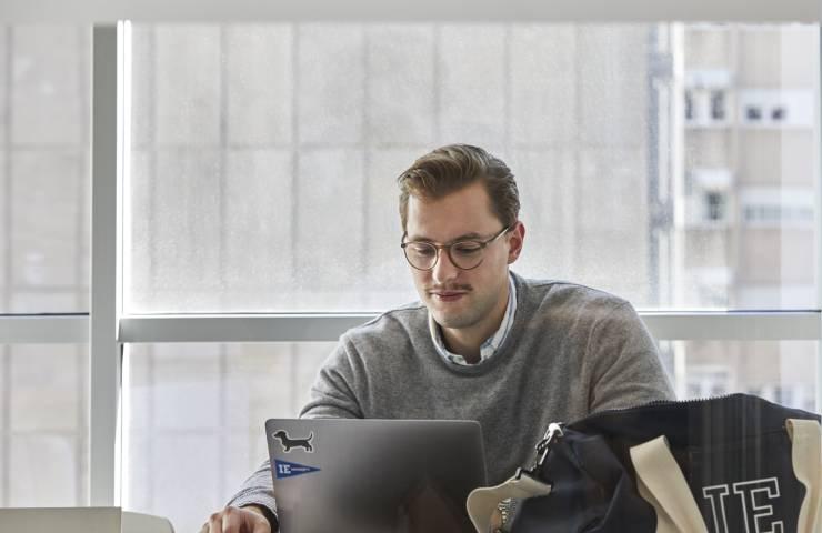 A man in glasses working on a laptop at a table in a brightly lit room with large windows.