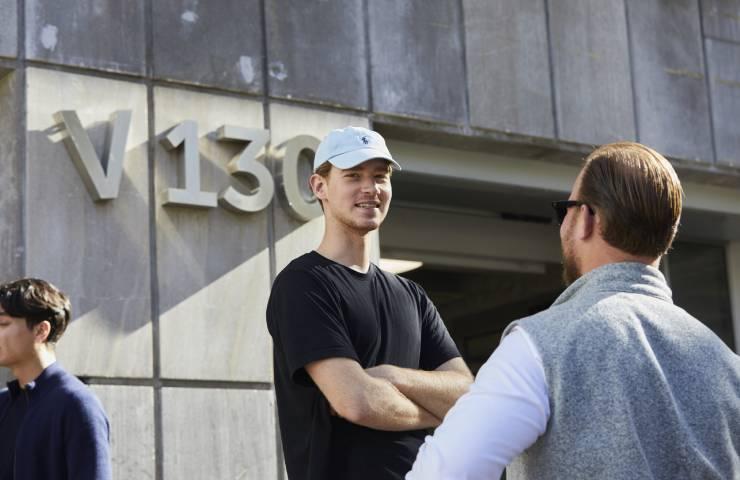 Two men engaging in a conversation in front of a building with the inscription 'V130'.