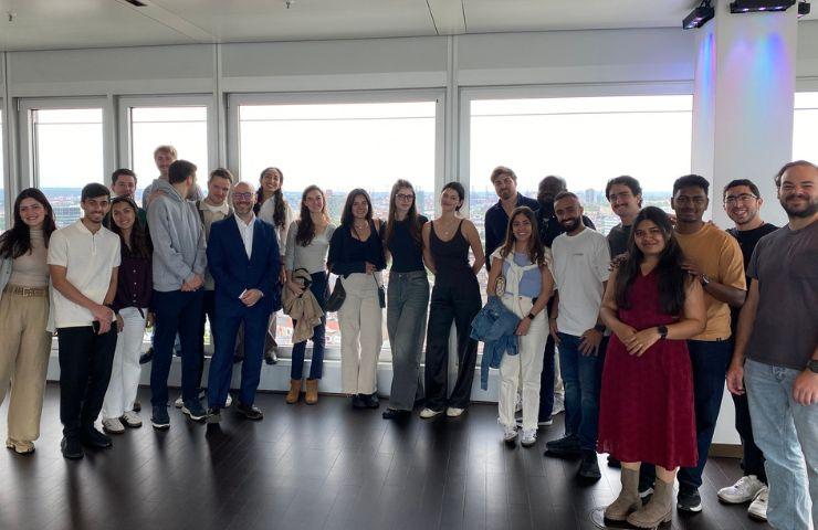 A diverse group of people standing together in a brightly lit room with large windows showing a city view.
