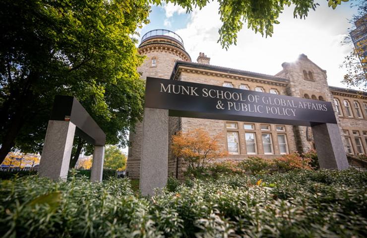 View of the Munk School of Global Affairs & Public Policy building behind a sign and lush greenery.