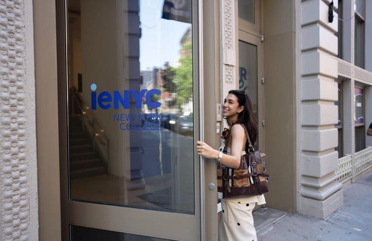 A young woman is smiling as she enters a building with a sign that reads 'ieNYC New York College'.