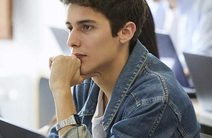 A young man in a denim jacket sits focused at a computer in a classroom setting, surrounded by other students also working on laptops.