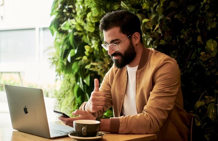 A man sitting at a table with a laptop and coffee, giving a thumbs up.