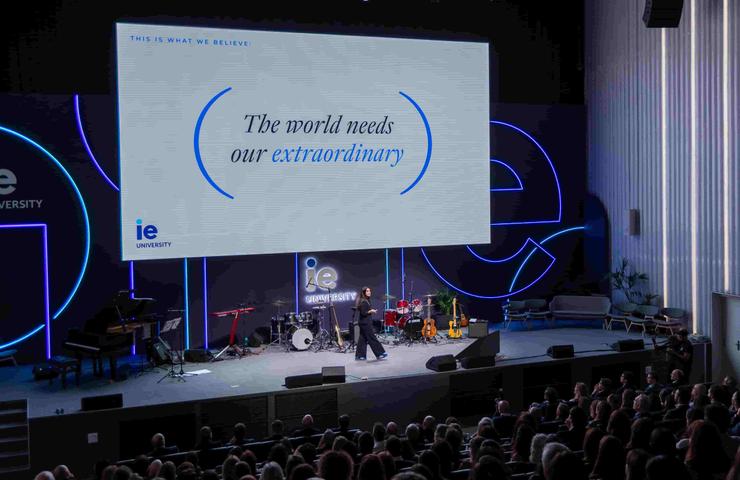 A speaker addressing an audience at an event, with a large screen displaying the message 'The world needs our extraordinary.'