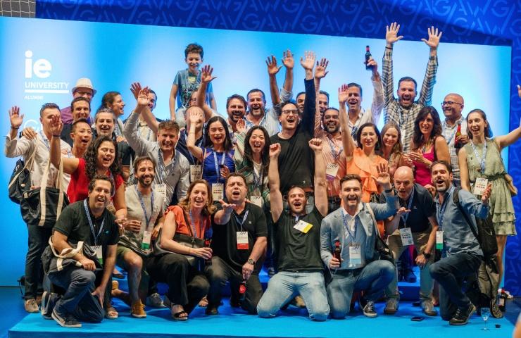 A group of people celebrating at a conference event with raised hands and cheerful expressions in front of a stage with 'IE University' branding.
