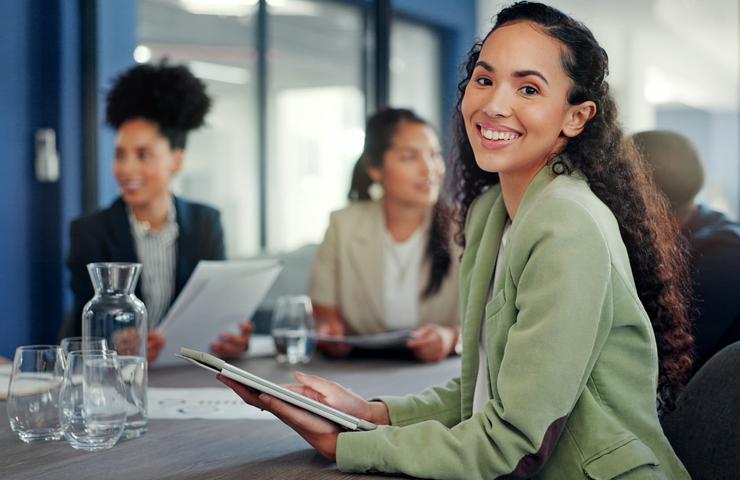 A woman in a green blazer smiles at a meeting table while colleagues engage in discussion.