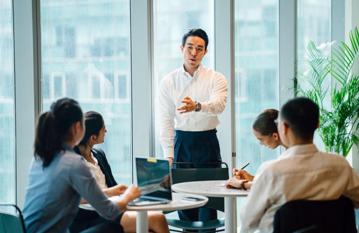 A man stands presenting to colleagues at a round table in a brightly lit office.