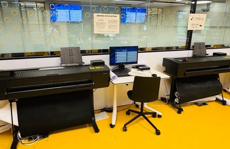 The image shows two large printers beside a computer and a chair in a bright workspace.