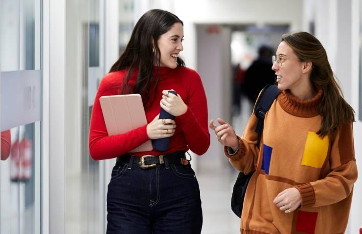 Two women walking and talking in a corridor, one holding a laptop and the other holding coffee cups.