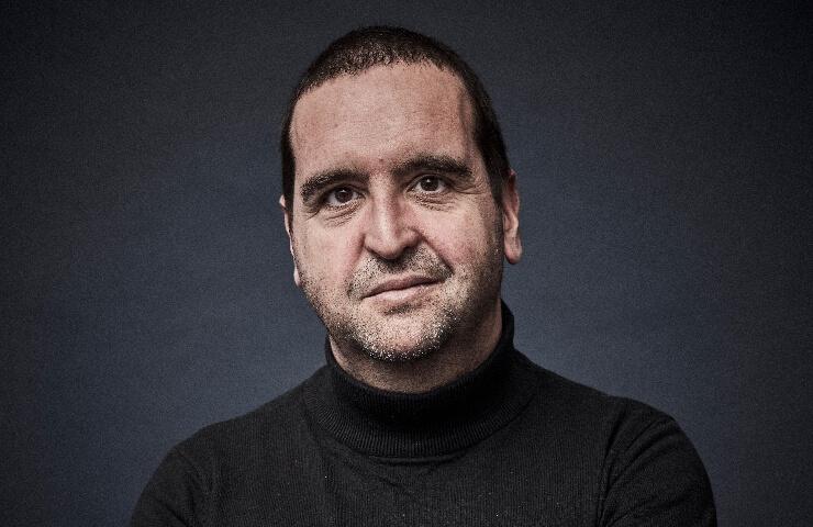 Portrait of a smiling man wearing a black turtleneck, set against a blue-grey background.