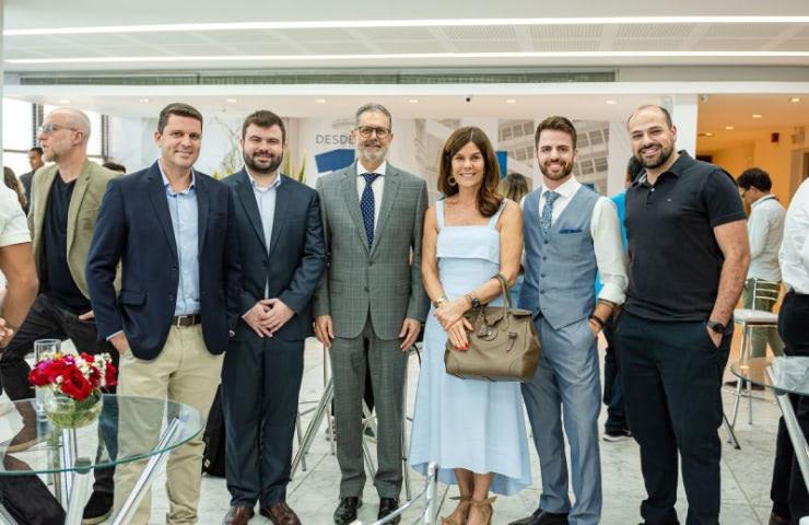 A group of six adults, three men and three women, pose for a photograph at an indoor event with other attendees in the background.