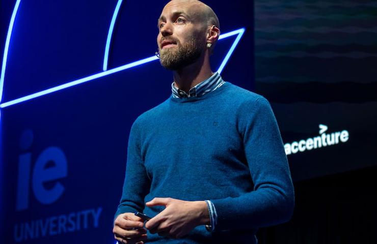 A man in a blue sweater is giving a presentation at a conference with a neon blue and purple graphical background.