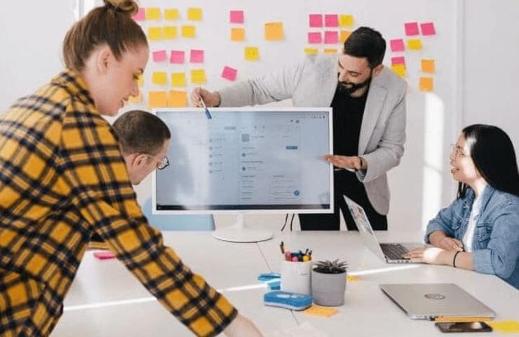 A group of colleagues collaboratively working around a digital monitor in a bright office decorated with sticky notes.
