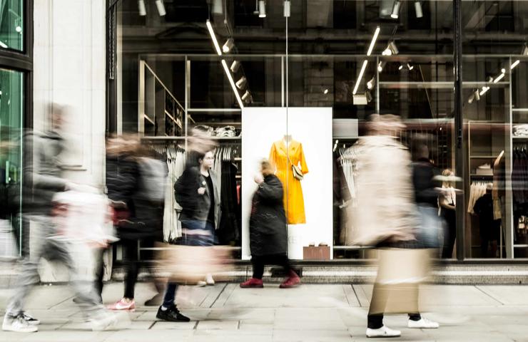 Blurred figures walking past a fashion store display with a bright yellow jacket on a mannequin.