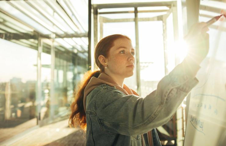 A young woman is writing on a glass board in a bright, sunlit workspace.