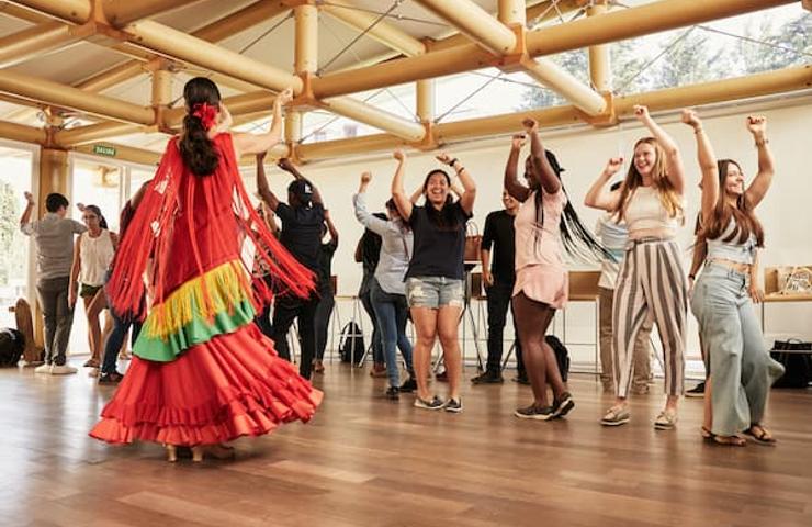 A diverse group of people are joyfully participating in a dance class led by a woman in a colorful traditional dress.