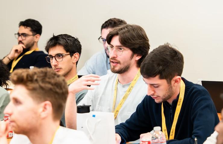 A group of young men engaged in a discussion at a conference, with one speaking and the others listening.