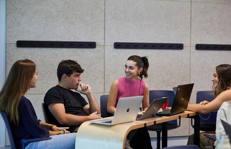 Four young adults are engaged in a discussion at a table with laptops in a modern classroom setting.