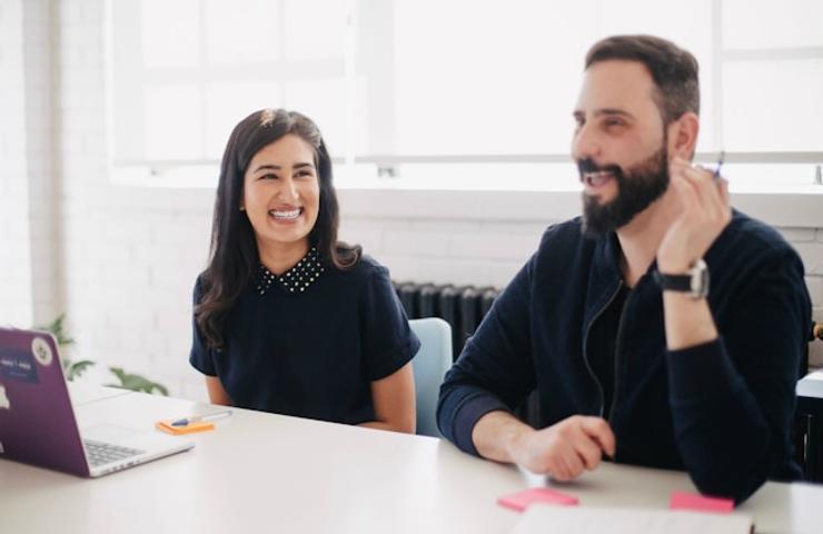 Two professionals, a man and a woman, smiling while sitting at a desk with a laptop in an office setting.