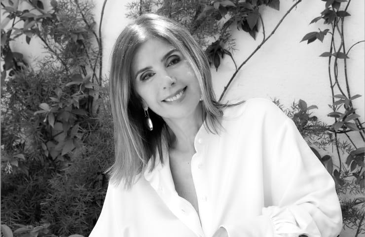 A black and white photo of a smiling woman in a white shirt standing against a background of green foliage.