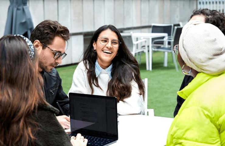 A group of young adults laugh and discuss while sitting around a table with a laptop outdoors.