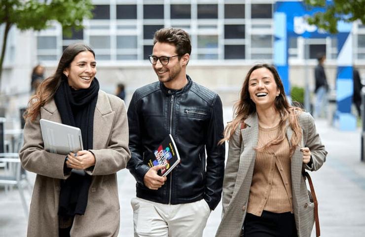 Three young adults walking and laughing together in an urban setting, with one holding a laptop and another holding books.