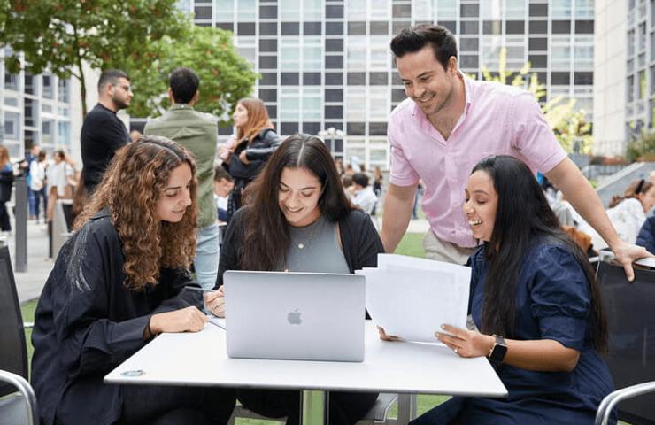 A group of four people, three women and one man, engaging with laptops and papers at a table outdoors.