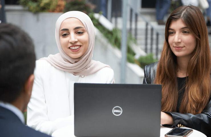 Two women, one wearing a hijab, are sitting around a table with a laptop, engaged in a conversation with a man whose back is to the camera.