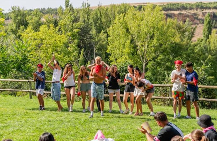 A group of people of various ages engaging in a playful activity outdoors in a grassy area with trees in the background.