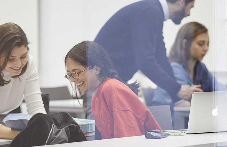 Two women smiling and looking at a laptop in a bright, busy office environment.