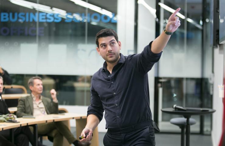 A man in a business casual outfit is presenting or lecturing to an audience in a modern office or classroom setting.