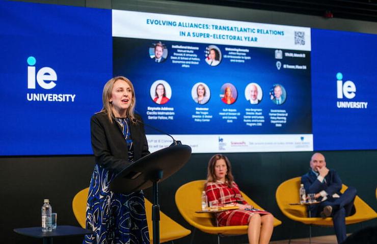 A woman is speaking at a podium during a panel discussion at an event related to transatlantic relations, with other panel members seated nearby and information displayed on a large screen above.