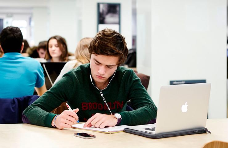A young man studying in a busy university library while listening to music on his headphones.