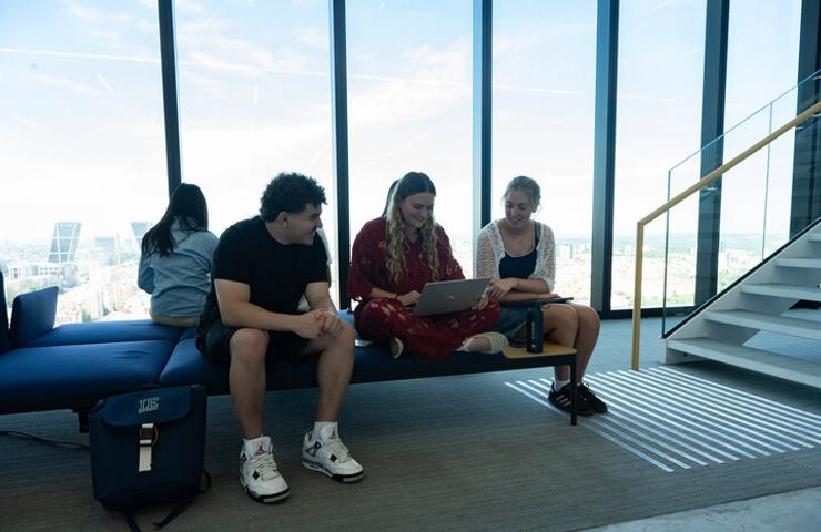 Three people are sitting on a bench near a large window, with one person working on a laptop.