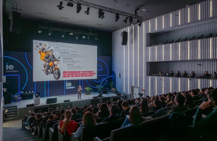 A speaker presents in front of a large audience at IE University, with a visual display featuring a motorcycle and text.