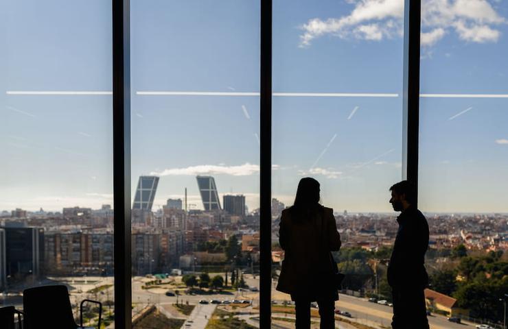 Two silhouetted figures are conversing in front of a large window showcasing a city skyline.